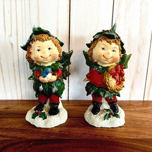 Peut inclure: Deux figurines de lutins de Noël. Chaque lutin a des cheveux bruns bouclés, un visage souriant et porte une tenue rouge et verte. L'un tient un oiseau bleu, l'autre une couronne. Les deux se tiennent sur une base enneigée.