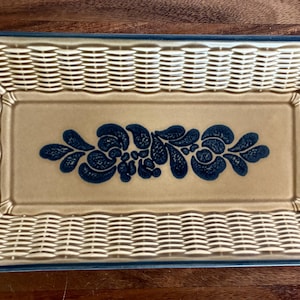 May include: A rectangular ceramic basket with a cream-colored base and a dark blue floral pattern in the center. The sides feature a molded basket-weave texture, and the entire rim is outlined in a dark blue border, creating a structured design.