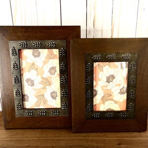 May include: Two wooden picture frames with dark brown frames and decorative metal accents. The frames hold floral print artwork with white flowers on a peach background. The frames are displayed on a wooden surface.