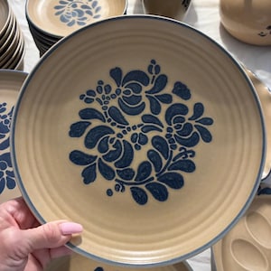 May include: A round ceramic dinner plate with a tan base and a dark blue floral pattern in the center. The plate features a series of concentric circular ridges along the rim and a dark blue band around the outer edge, matching the central design.