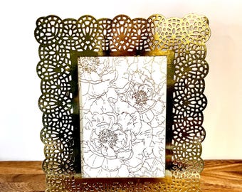 May include: A gold-colored, rectangular picture frame with an ornate, openwork design. The frame holds a print with a floral pattern in black and white. The frame is made of metal and has a decorative, scalloped edge.