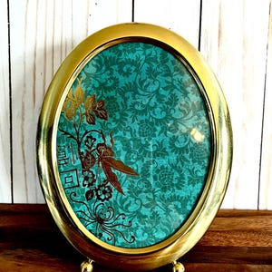 May include: Oval brass picture frame with a teal floral background and gold accents. The frame has a decorative design with a floral motif. The frame is supported by small, round feet and is displayed on a wooden surface.