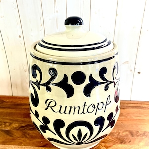 May include: A cream-colored ceramic crock with a matching lid, featuring dark blue hand-painted floral and scroll patterns. The word Rumtopf is written in black script on the front. The vessel has a rounded body and a small knob on the lid.