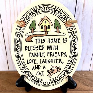 Könnte beinhalten: Eine weiße ovale Keramikplakette mit schwarzem Rand und einem dekorativen Design. Die Plakette zeigt eine Comic-Illustration eines Hauses mit Bäumen, einem Weg und einer Katze. Der Text auf der Plakette lautet: "This home is blessed with family, friends, love, laughter, and a cat."