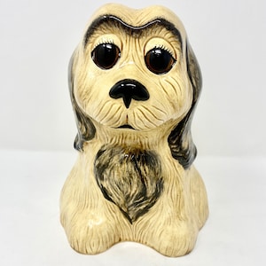 Op de afbeelding: Een keramiek beeldje van een hond met grote bruine ogen, een zwarte neus en bruin en crèmekleurig haar. De hond zit met zijn hoofd lichtjes gekanteld.