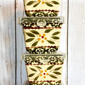 May include: Four stacked ceramic food storage containers with clear plastic lids. The containers have a cream-colored base with a floral design in green, yellow, and red. The lids are clear plastic.