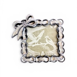 Puede incluir: Un marco de fotos cuadrado plateado con un lazo en la esquina superior izquierda. El marco tiene un borde decorativo y contiene una impresión de dos pájaros en una rama. El lazo está adornado con pedrería.