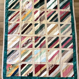 May include: A rectangular quilt featuring a grid of square blocks, each composed of diagonal fabric strips in various colors and patterns. The blocks are separated by beige sashing and framed by a dark green border with a light beige outer edge.