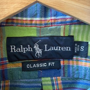 Puede incluir: Primer plano de una camisa Ralph Lauren con un estampado de cuadros en azul, verde y naranja. La etiqueta negra muestra el nombre y el logotipo de la marca, con "CLASSIC FIT" debajo. La etiqueta de talla indica "S".