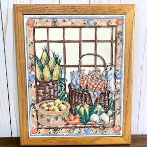 May include: A framed ceramic tile with a country kitchen theme. The tile features a window with a checkered pattern, a basket of corn, a basket of fruit, and a bowl of lemons. The tile is surrounded by a border of hearts and flowers.