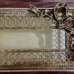 May include: A rectangular gold-toned metal tissue box cover featuring an intricate lattice pattern along the sides and a prominent decorative bow attached to the top right corner. The surface has a textured finish with detailed border engravings.