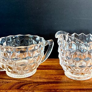 May include: A clear glass sugar bowl and creamer set. Both pieces feature a diamond-cut design and curved handles. The sugar bowl is round, and the creamer has a spout. The set is displayed on a wooden surface.