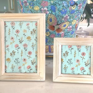 Puede incluir: Dos marcos de fotos de madera blanca con un patrón floral sobre un fondo azul claro. El marco más grande es vertical y el marco más pequeño es horizontal.