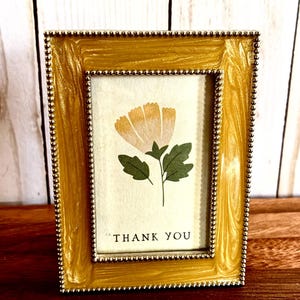 May include: A gold-colored picture frame with a floral print and the words "THANK YOU". The frame has a beaded border and a glossy finish. The floral print features a yellow flower with green leaves.
