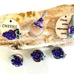 May include: A collection of wine-themed decorative items. Includes a bottle, a tag with "CHEERS", and a tag with "Cheap wine, expensive cork". The items feature purple grape clusters and silver accents. The items are displayed on a light brown, textured surface.