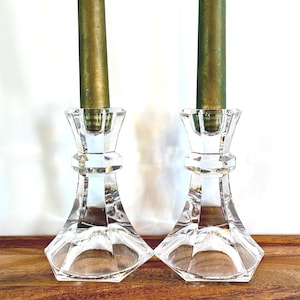 Puede incluir: Dos candelabros de cristal transparente facetado con velas doradas. Los candelabros tienen un diseño geométrico y están sobre una superficie de madera. El fondo es blanco.
