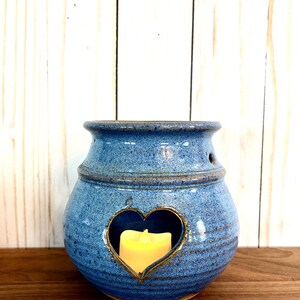May include: A blue ceramic candle holder with a heart-shaped cutout. The holder has a textured surface and a small hole on the side. A yellow candle is inside the holder, creating a warm glow. The candle holder is a decorative item.