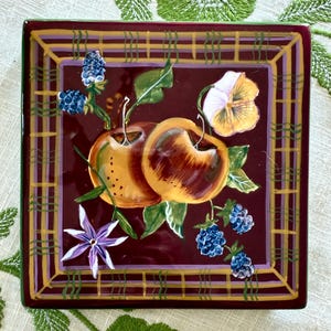 Puede incluir: Plato decorativo cuadrado de cerámica con fondo granate y diseño pintado de dos manzanas, moras y una flor. El plato tiene un borde dorado y morado. El plato mide aproximadamente 20 cm cuadrados.