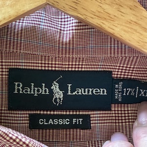May include: A red and white check button-down shirt with the Ralph Lauren logo. The shirt is labelled "Classic Fit" and is size 17 1/2 XL. The shirt is hanging on a wooden hanger.