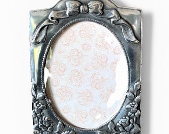 May include: Silver-toned oval picture frame with a bow at the top and floral detailing. The frame has an oval opening and a patterned background with pink roses. The frame is made of metal.