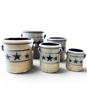 Puede incluir: Un conjunto de cinco jarrones de gres de diferentes tamaños. Cada jarrón tiene una base beige con diseños de estrellas y rayas azules. Los bordes y el interior son de color azul oscuro, y cada uno tiene dos asas pequeñas.