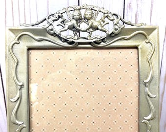 May include: A silver picture frame with an ornate design featuring two cherubs. The frame has a light brown mat with a white polka dot pattern.