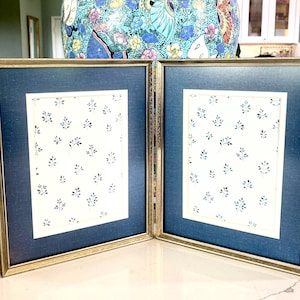 Puede incluir: Un marco de fotos de doble apertura con un marco dorado. Cada apertura tiene un passe-partout azul y un fondo blanco con un pequeño patrón floral azul.