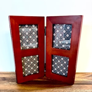 Può includere: Una cornice in legno rosso ciliegia con quattro aperture rettangolari. Ogni apertura mostra un inserto a fantasia con disegni floreali bianchi su sfondo scuro. La cornice è incernierata e può stare in piedi.