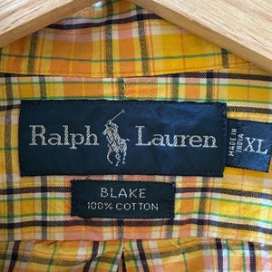 May include: Close-up of a yellow, orange, and green plaid shirt with the Ralph Lauren logo and "BLAKE 100% COTTON" label. The shirt is size XL and made in India. The shirt is hanging on a wooden hanger.