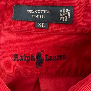 Puede incluir: Primer plano de una camisa de pana roja. La etiqueta dice "100% Algodón" y "XL". La camisa presenta el logotipo "Ralph Lauren" en escritura negra. La etiqueta también indica que la camisa fue fabricada en las Islas Marianas del Norte.