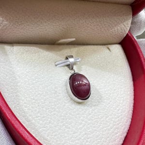 May include: A silver pendant featuring a smooth, oval-shaped deep red gemstone with a visible star-like light reflection. The pendant is held by a small silver bail and is displayed inside a heart-shaped jewelry box with a cream-colored interior.