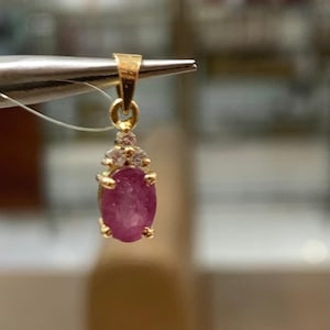 Peut inclure: Un pendentif en or avec une grosse pierre précieuse ovale en rubis. Le rubis est d'un rose profond et est entouré de petites pierres précieuses claires. Le pendentif est tenu par une pince.