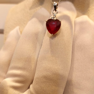 May include: A heart-shaped red gemstone pendant set in a silver-toned metal frame. The pendant features a small, clear accent stone at the top, positioned just below the silver bail, which is attached to a thin, clear monofilament hanging cord.