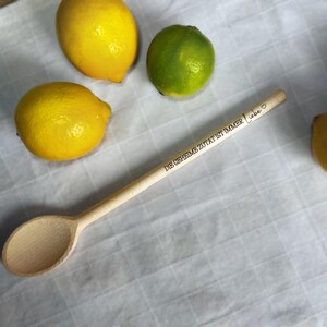 Könnte beinhalten: Ein Holzlöffel mit dem Text "DIE GEHEIME ZUTAT IST IMMER Liebe" eingraviert. Der Löffel ist von Zitronen und einer Limette umgeben.