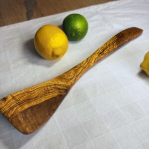 Könnte beinhalten: Ein Holzspatel mit einer natürlichen Holzmaserung. Der Spatel hat einen langen Stiel mit dem eingravierten Text "DIE GESCHICHTE DES HOLZES". Der Spatel liegt auf einer weißen Oberfläche mit Zitronen und einer Limette.