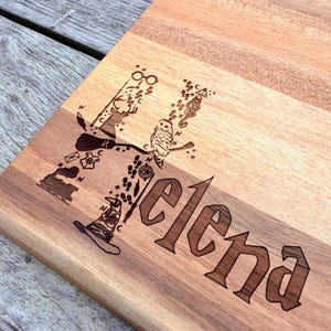 Könnte beinhalten: Ein Schneidebrett aus Holz mit einem Harry-Potter-Motiv. Das Brett ist mit dem Namen "Elena" graviert und zeigt verschiedene Harry-Potter-Symbole, darunter ein Hogwarts-Wappen, einen goldenen Schnatz und eine Brille.
