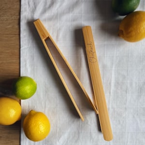 Könnte beinhalten: Ein Paar natürliche Bambuszangen mit dem Wort "Chefkoch" auf einer Seite graviert. Die Zangen liegen auf einer weißen Leinenoberfläche mit Zitronen und Limetten.
