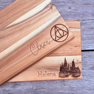 Könnte beinhalten: Zwei Holzschneidebretter mit eingravierten Designs. Ein Brett zeigt den Namen "Clara" und ein geometrisches Symbol. Das andere Brett hat den Namen "Helena" und eine Schlossillustration. Beide Bretter sind aus hellem Holz gefertigt.