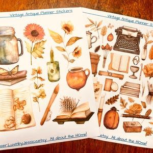 May include: Two sheets of planner stickers featuring watercolor illustrations in warm orange and brown tones. Designs include a mason jar, sunflower, typewriter, books, candle, pottery, pinecone, and leaves. Text reads Vintage Antique Planner Stickers.