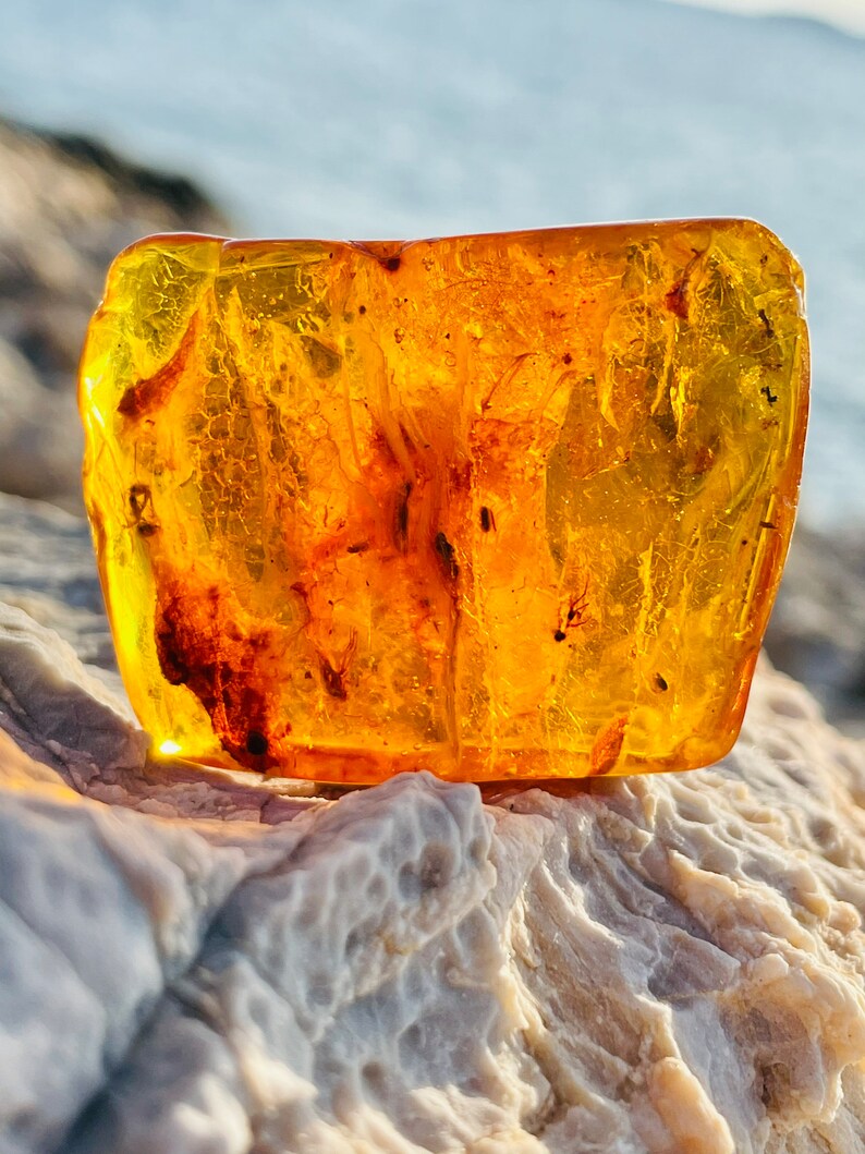 May include: A large, amber-colored piece of fossilized tree resin with inclusions of insects. The amber is translucent and has a warm, golden hue. It is resting on a light gray, textured surface.