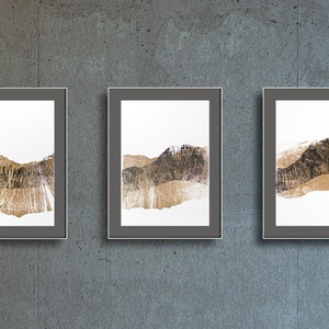 May include: Three framed abstract art prints. Each print features a mountain landscape design in shades of gold, brown, and white. The artwork is set against a white background and framed in a dark gray frame. The prints are displayed on a gray concrete wall.