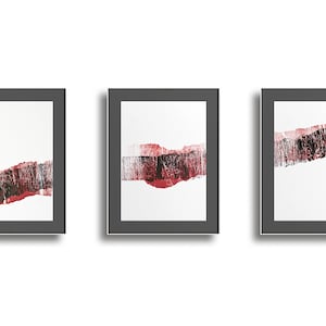May include: Three framed abstract art prints. Each print features a white background with a horizontal band of red and black textured brushstrokes. The frames are dark gray, and the overall style is modern.