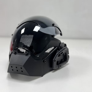 May include: A glossy black helmet with a visor and a geometric face guard. The helmet has a mesh vent and a handle on the side. The design is futuristic and sleek.