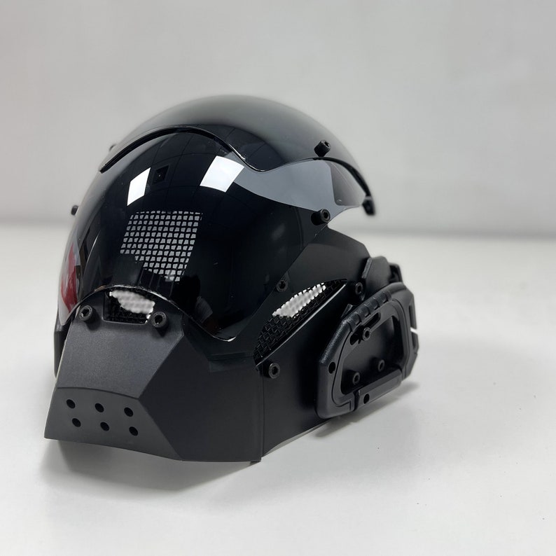 May include: A glossy black helmet with a visor and a geometric face guard. The helmet has a mesh vent and a handle on the side. The design is futuristic and sleek.