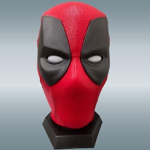 May include: A red and black Deadpool mask on a black stand. The mask has a textured surface and white eyes.