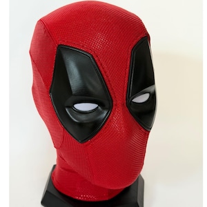 May include: A full-head mask featuring a vibrant red textured fabric exterior. The mask includes large, angular black eye frames with white mesh lenses, creating a distinct facial appearance. The design covers the entire head and neck area.