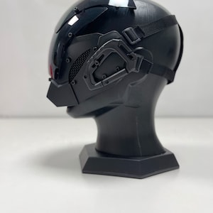 May include: A black futuristic helmet with a visor, mesh detailing, and adjustable straps. The helmet is mounted on a black display stand, showcasing its design and features. The overall aesthetic suggests a sci-fi or cosplay theme.