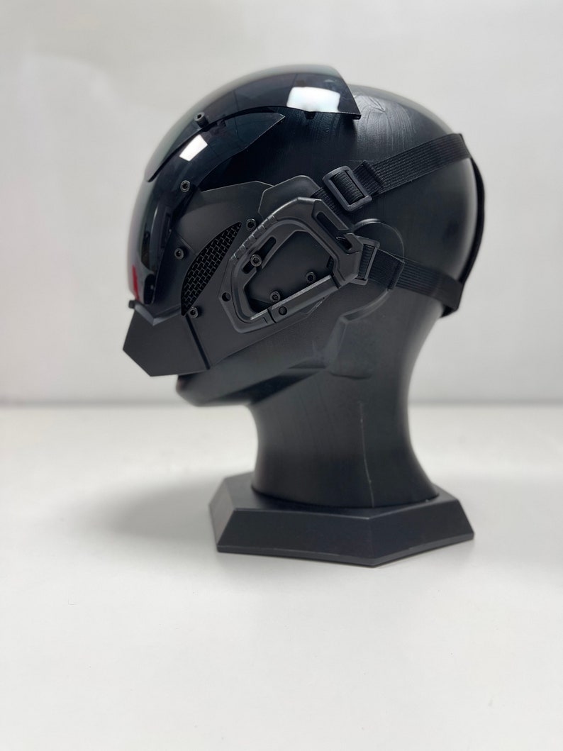 May include: A black futuristic helmet with a visor, mesh detailing, and adjustable straps. The helmet is mounted on a black display stand, showcasing its design and features. The overall aesthetic suggests a sci-fi or cosplay theme.