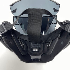 May include: Black tactical mask with tinted lenses and adjustable straps. The mask features a mesh vent and padded interior for comfort. The design is angular and modern, suitable for various activities.