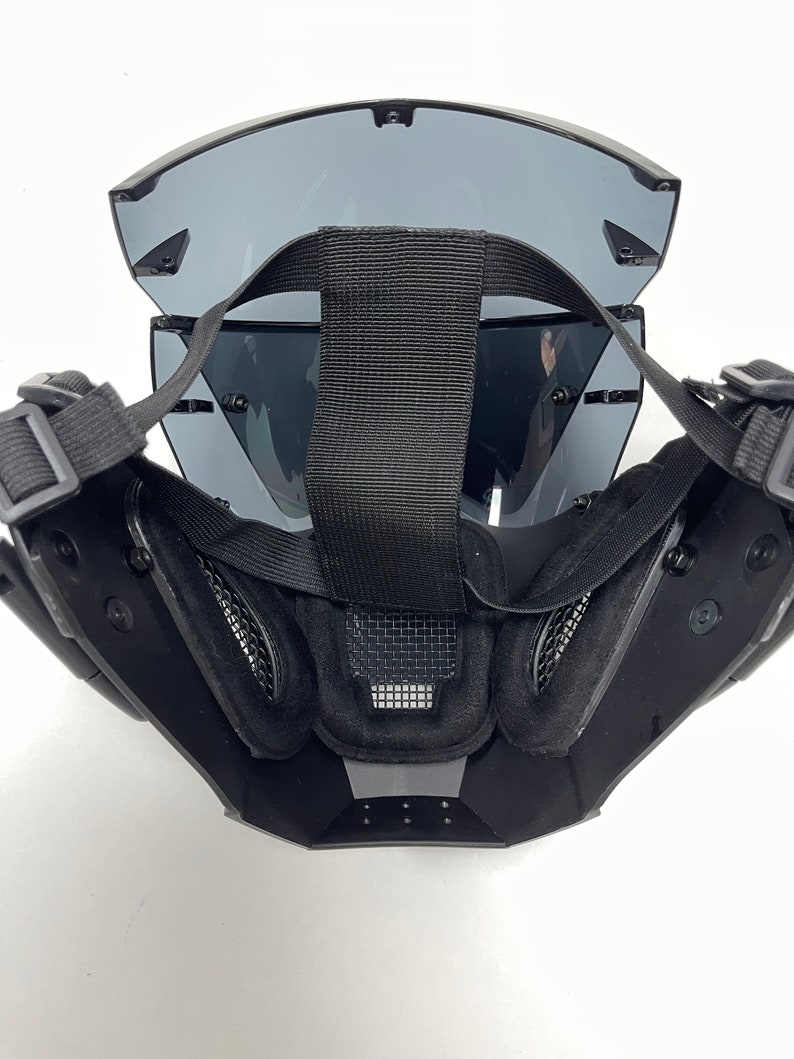 May include: Black tactical mask with tinted lenses and adjustable straps. The mask features a mesh vent and padded interior for comfort. The design is angular and modern, suitable for various activities.